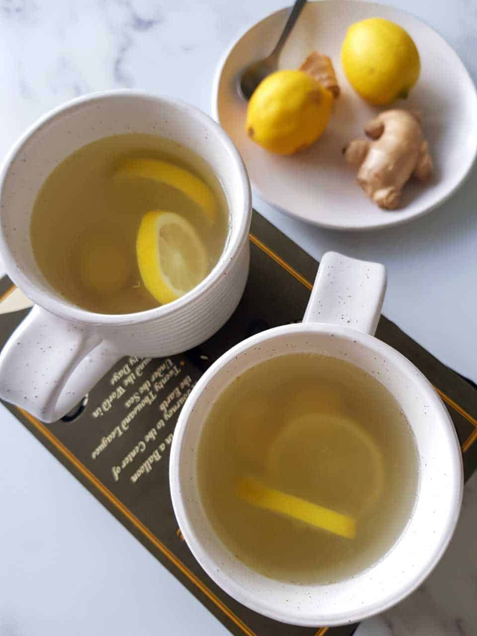 Hot lemon and ginger drink in cups on top of a book on a marble table.