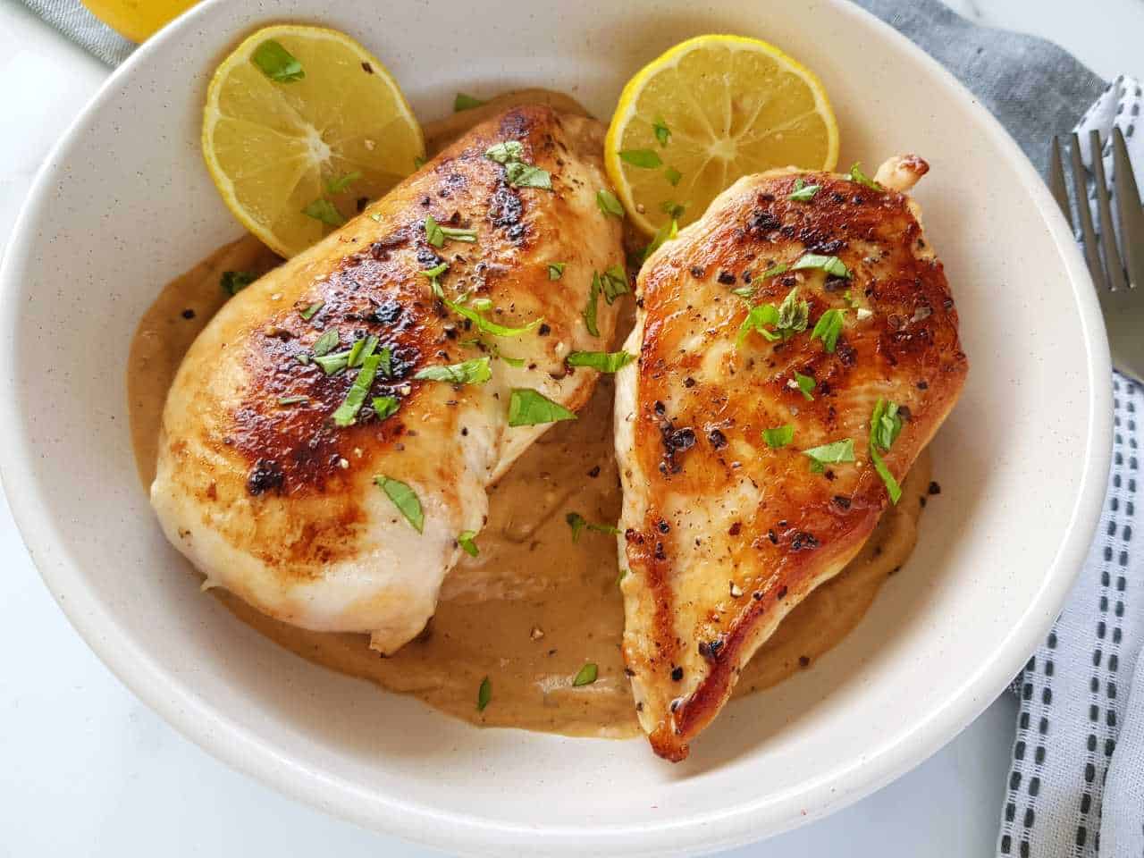 Lemon pepper chicken on a white plate with cutlery and lemon slices on the side.