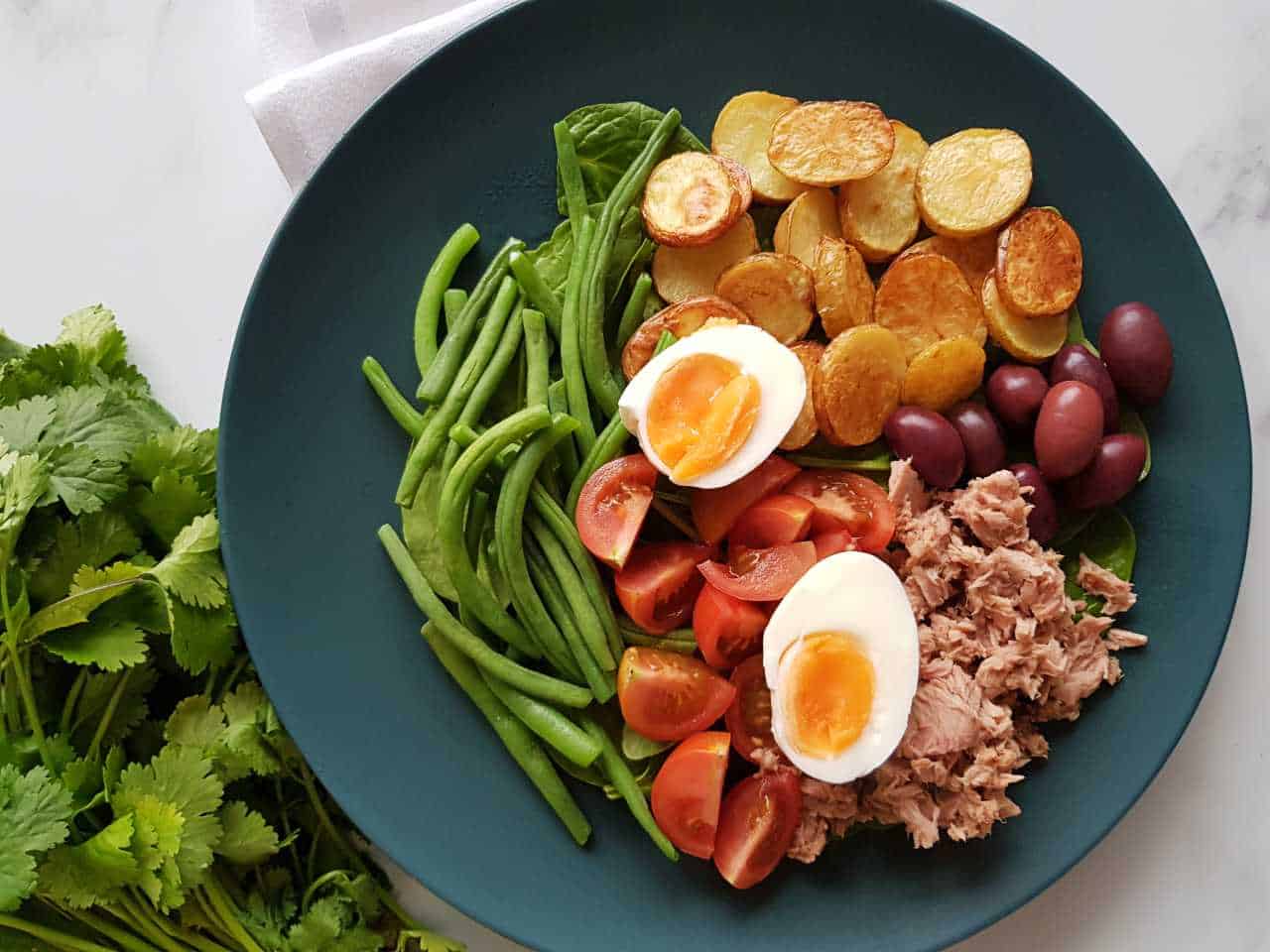 Nicoise salad on a blue plate on a marble table with fresh herbs on the side.