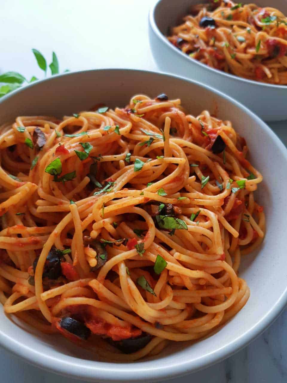 Pasta puttanesca garnished with fresh basil in white bowls on a white table.