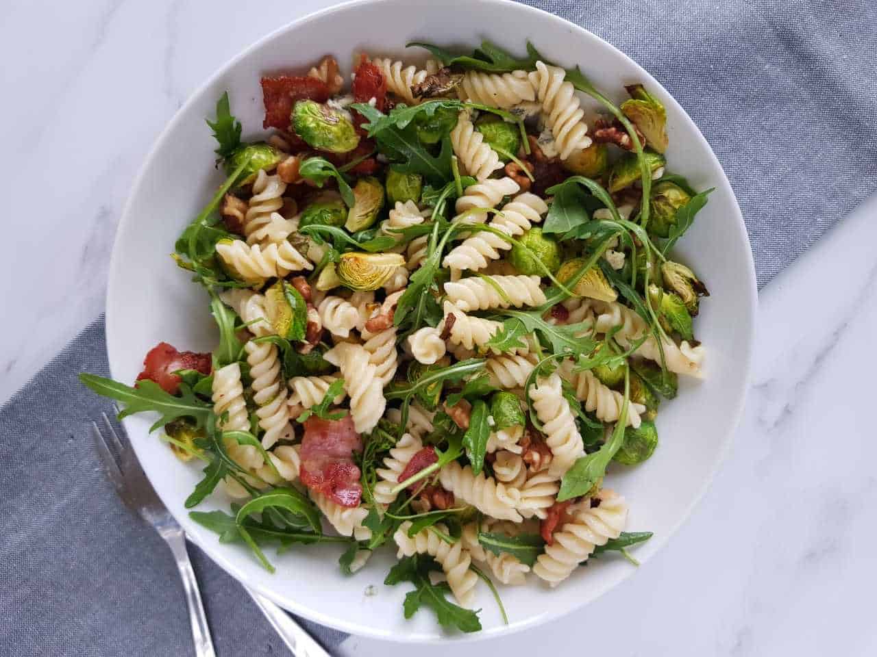 Pasta salad with brussels sprouts and bacon in a white bowl with forks and a grey tea towel on the side.