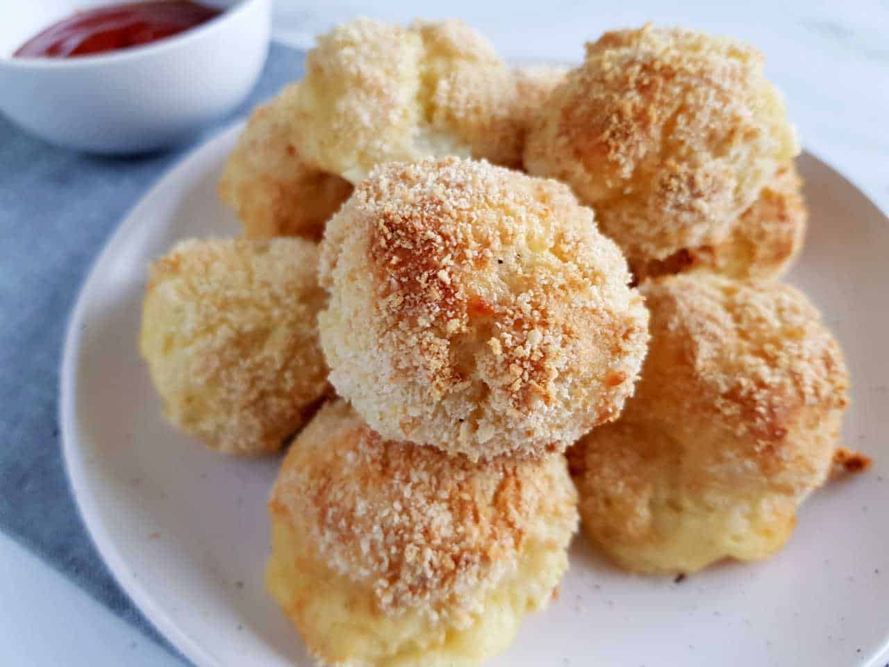 Potato and cheese balls on a white plate on a marble table with ketchup on the side.