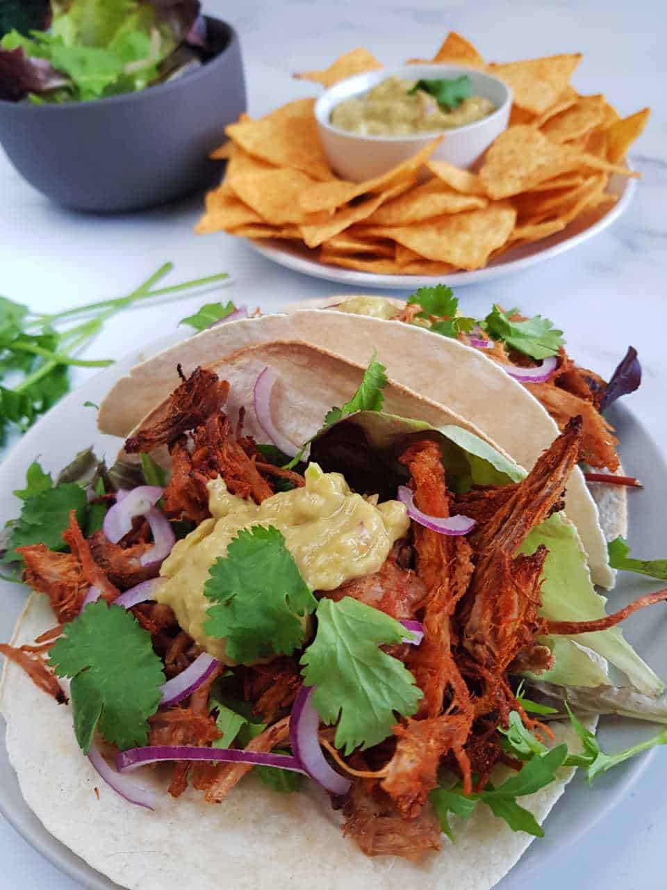 Slow cooker pulled pork tacos on a marble table with a plate of nachos in the background.