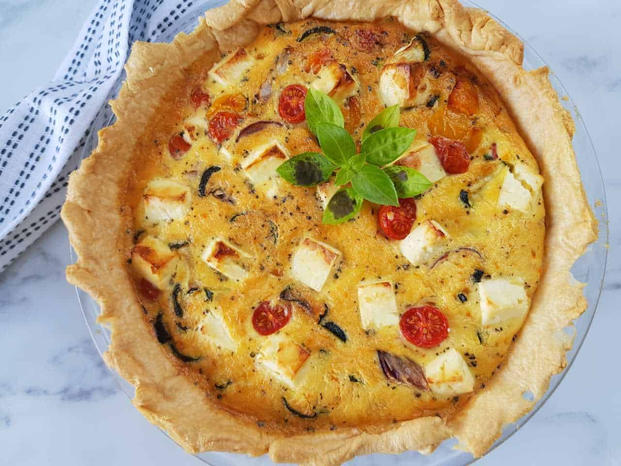 Quiche with feta and roasted vegetables on a marble table.
