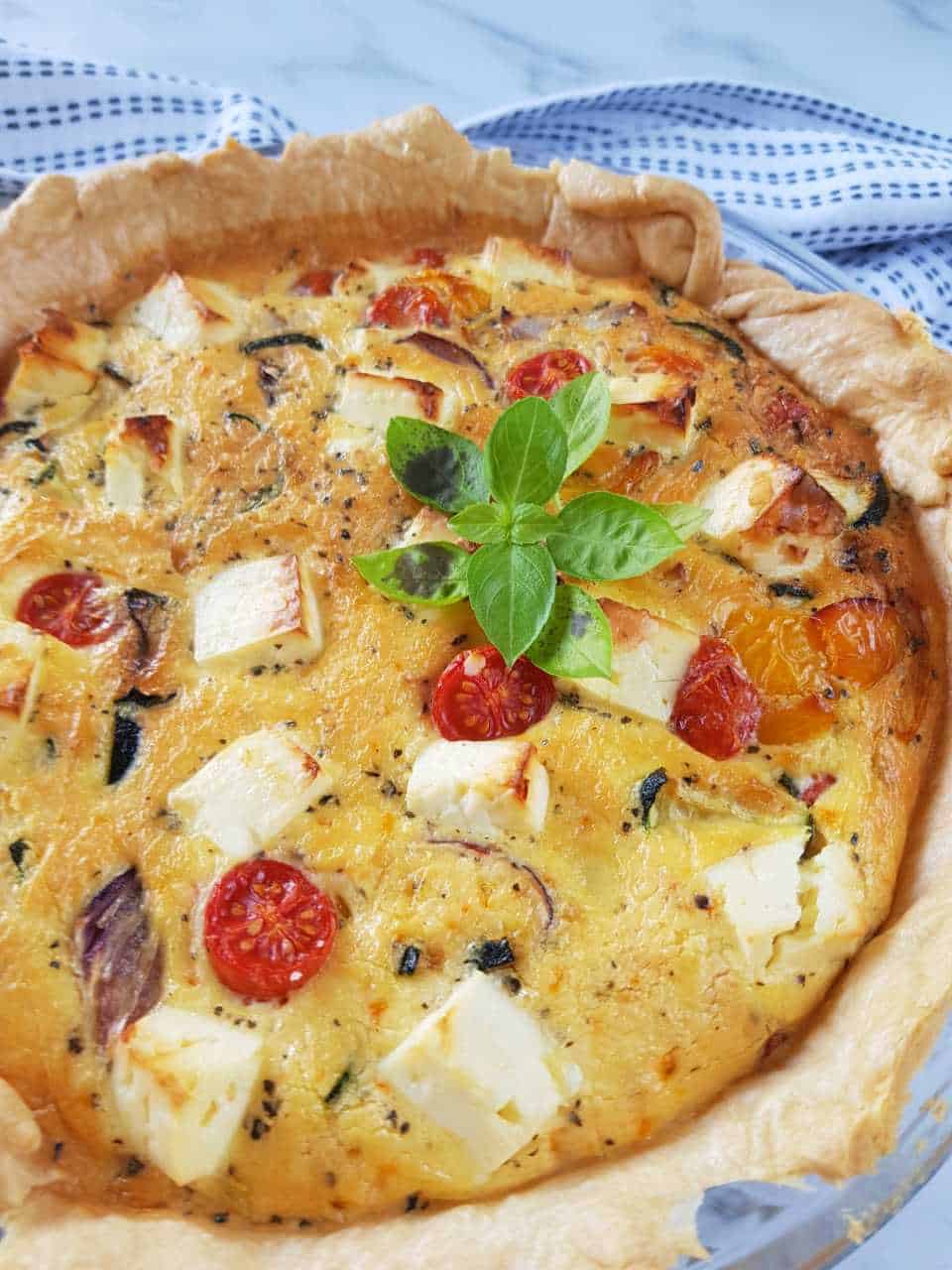 A quiche with cubes of feta and halves of cherry tomatoes. A sprig of basil is garnishing the top.