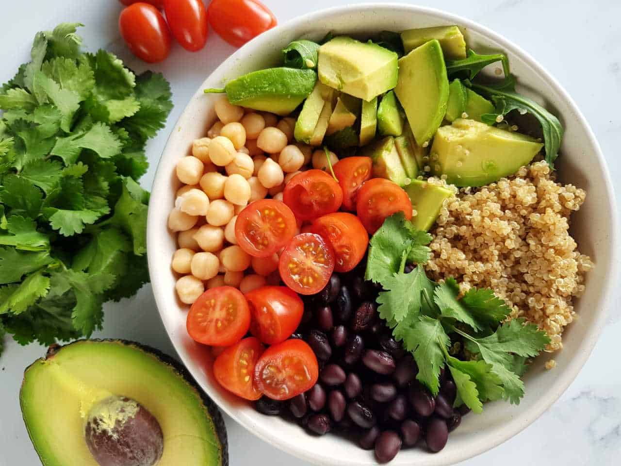 Quinoa salad with black beans, chickpeas and avocado in a bowl on a marble table, surrounded by tomatoes, coriander and avocado.