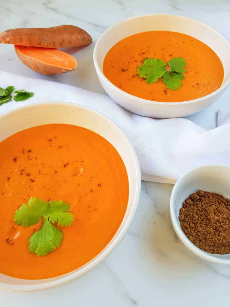 Red pepper and sweet potato soup in white bowls on a white table with a bowl of spices on the side.