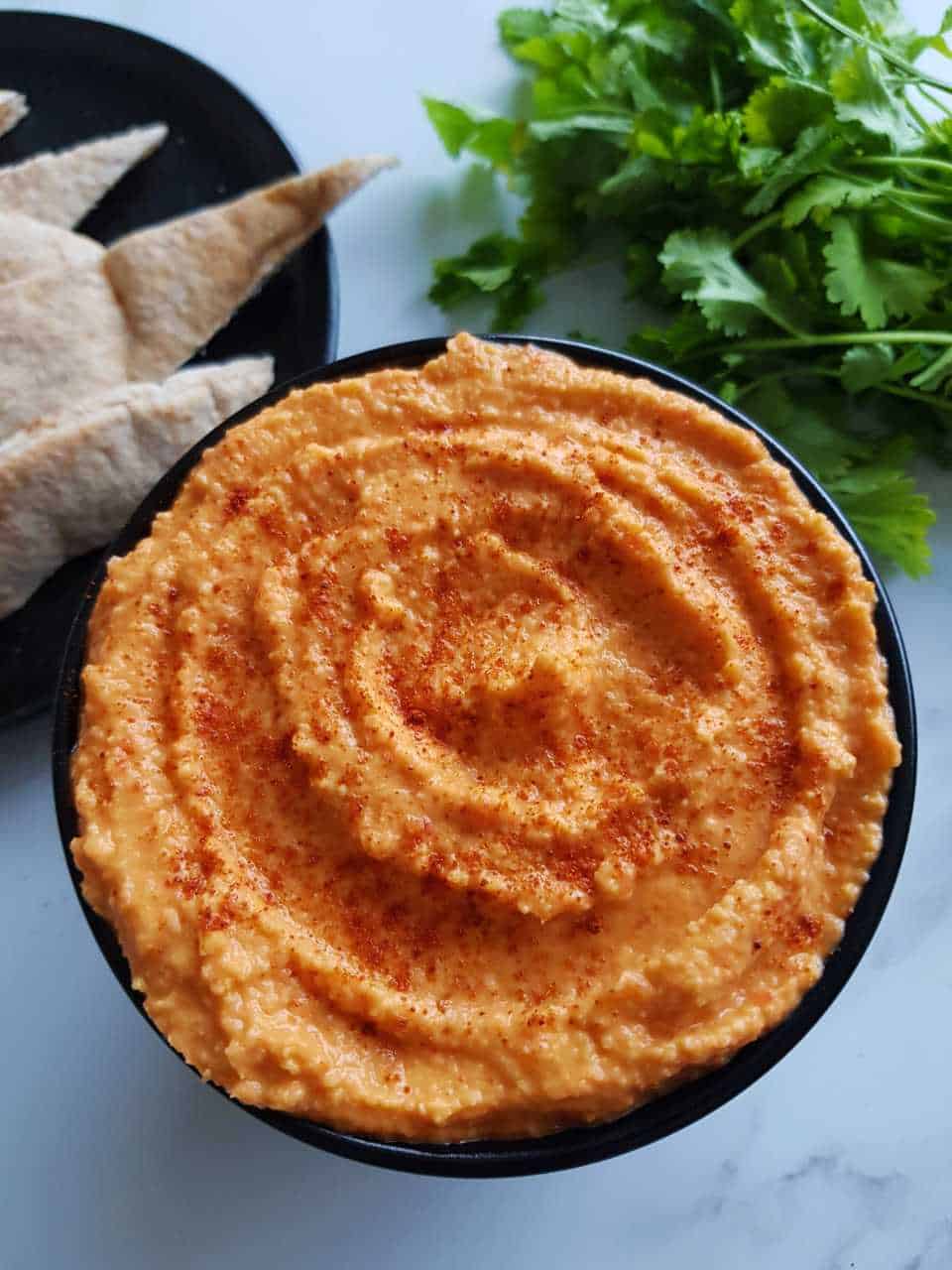 Red pepper hummus on a white table with cilantro and pita bread in the background.