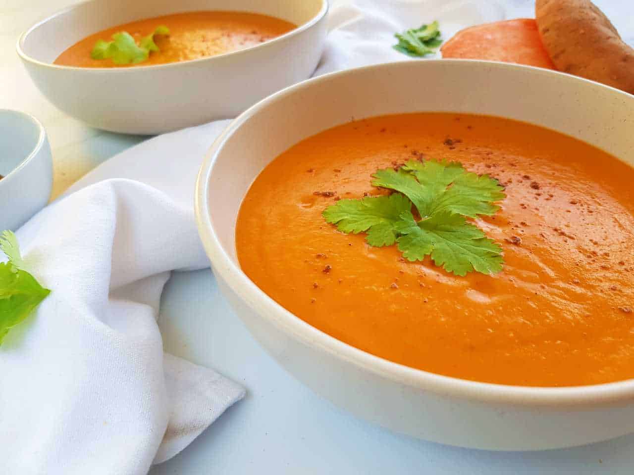 Roasted red pepper and sweet potato soup in white bowls with a white napkin and a sweet potato in the background.