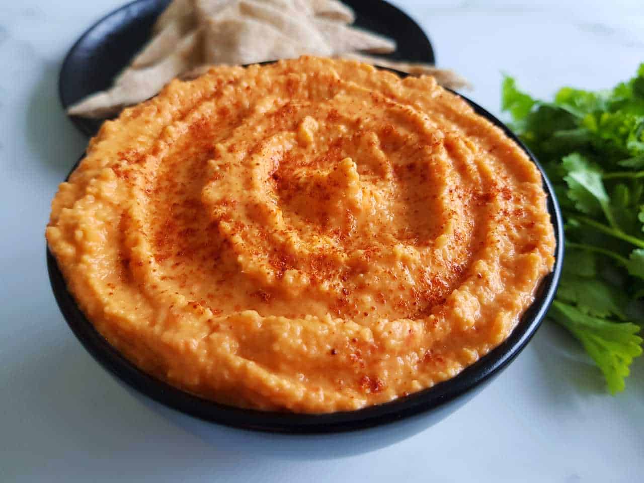 Roasted red pepper hummus in a black bowl with cilantro and pita breads in the background.