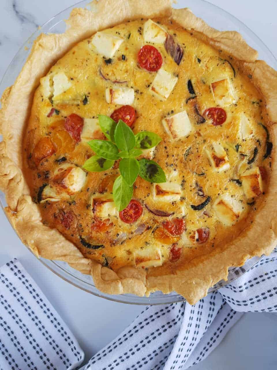 Roasted vegetable and feta quiche on a marble table with a tablecloth on the side.