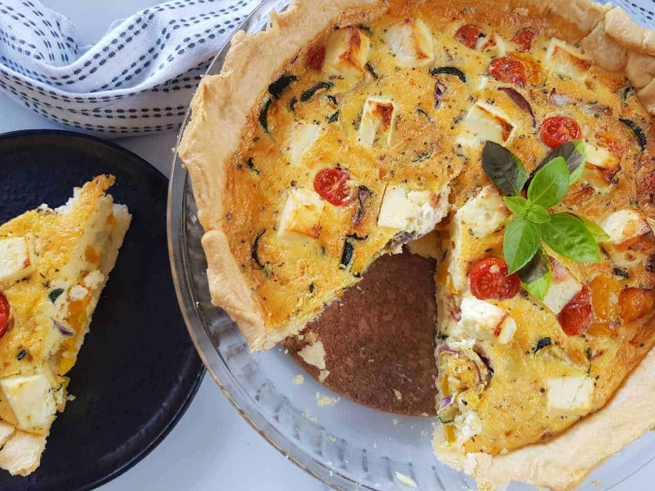 Roasted vegetable quiche with feta on a table, with a slice plated on the side.