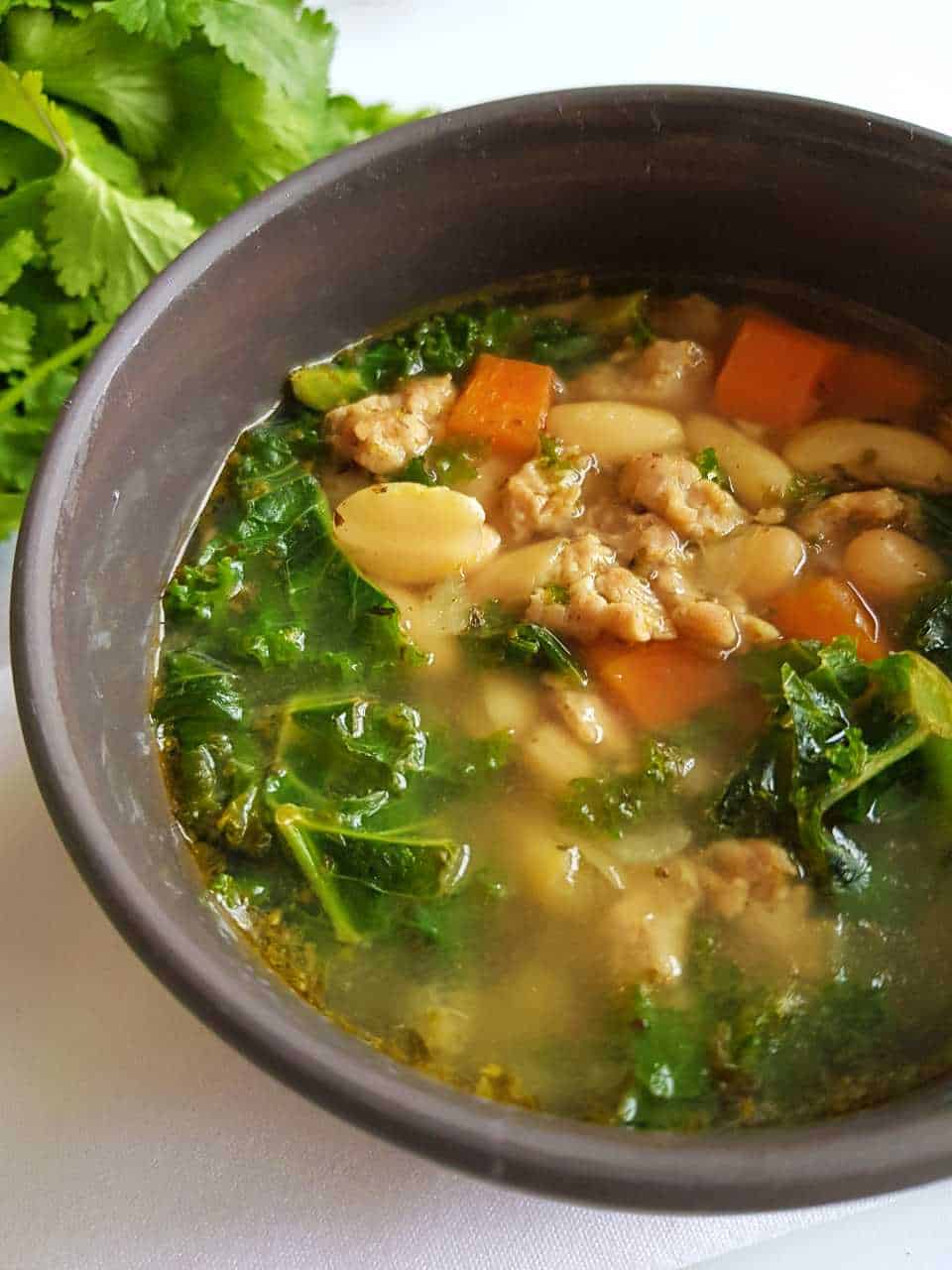 Sausage soup with vegetables in a grey bowl with fresh herbs in the background.