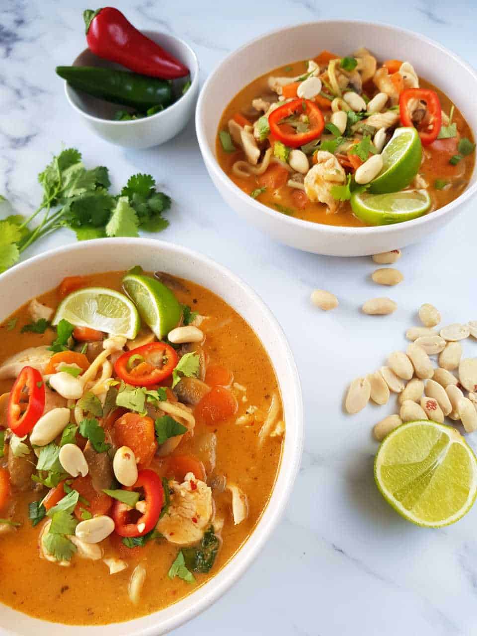 Thai chicken noodle soup in white bowls on a marble table with peanuts, lime, fresh coriander and chili scattered.