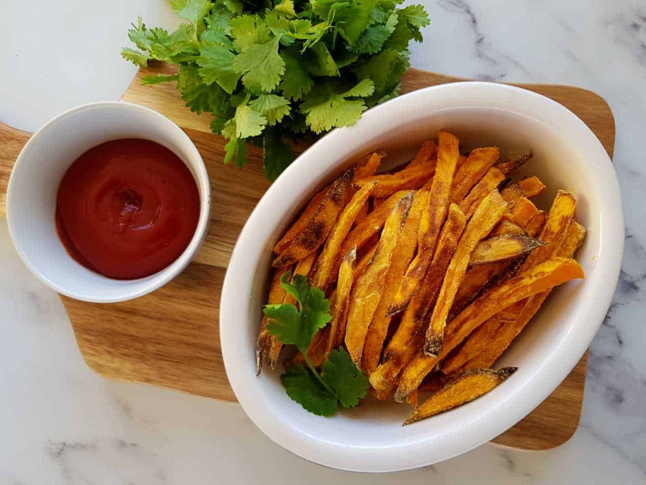 sweet potato chips in a white bowl with ketchup and fresh herbs on the side.