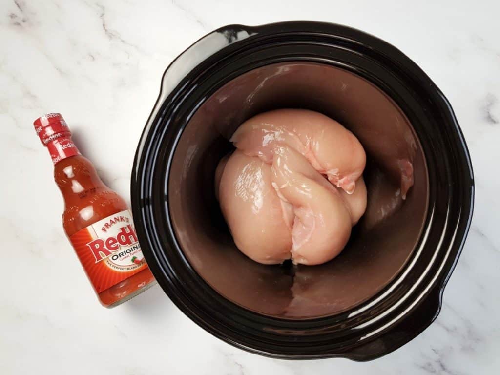 Raw chicken breast fillets in a slow cooker with buffalo sauce on the side.