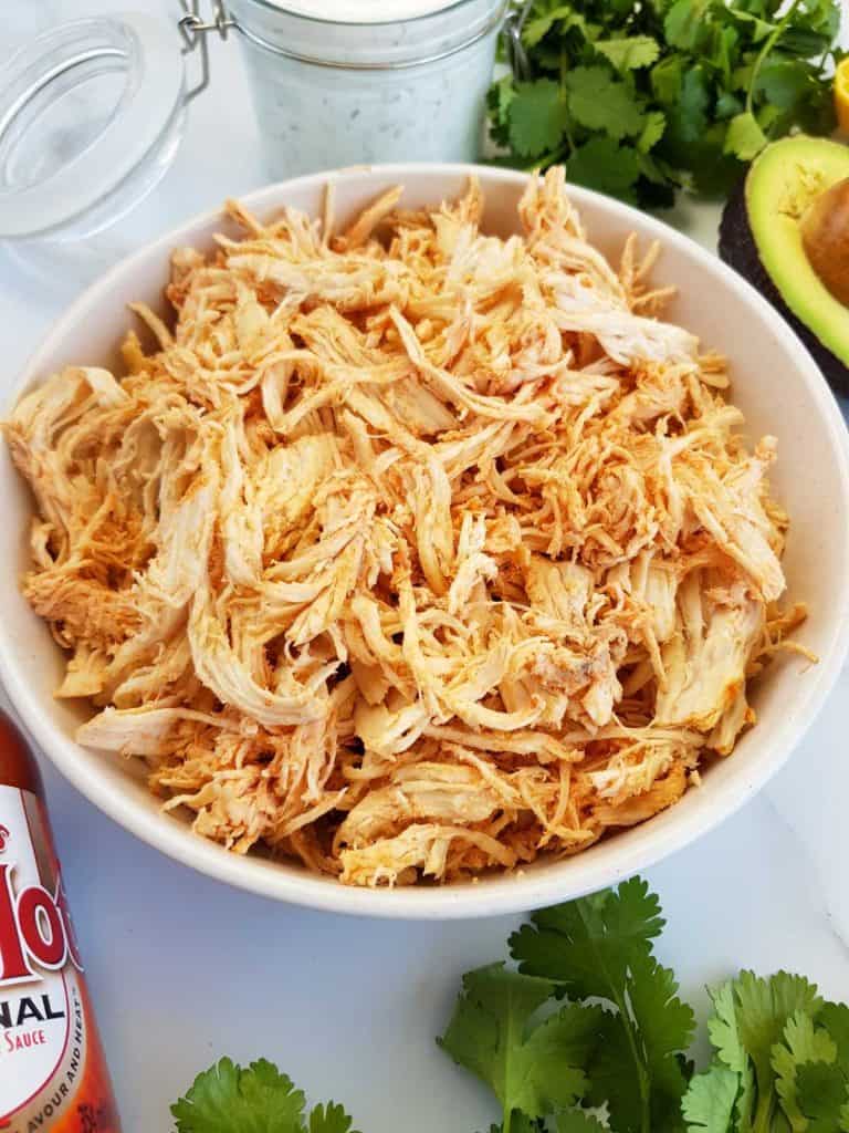 Shredded buffalo chicken in a bowl with buffalo sauce, ranch, avocado and cilantro on the side.