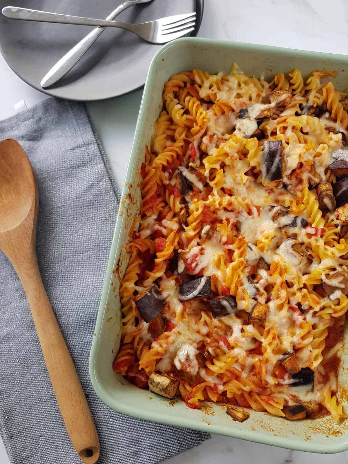 Eggplant and mozzarella pasta bake in a baking dish next to a plate with cutlery, a tea towel and a wooden serving spoon.