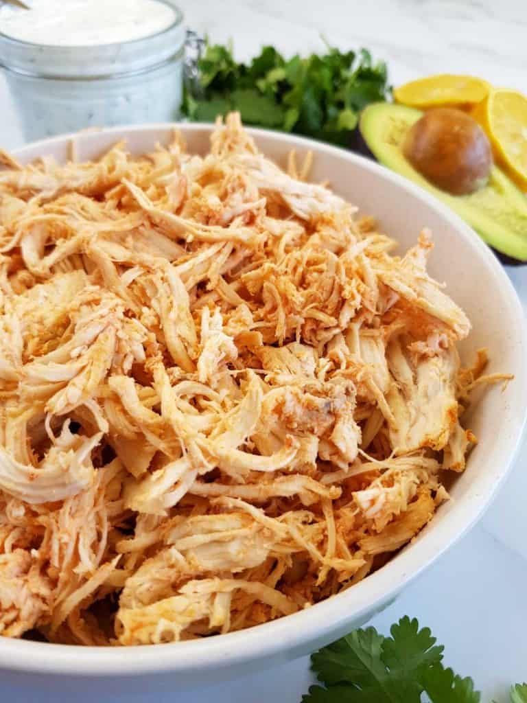 Shredded buffalo chicken in a bowl with buffalo sauce, ranch, avocado and cilantro on the side.