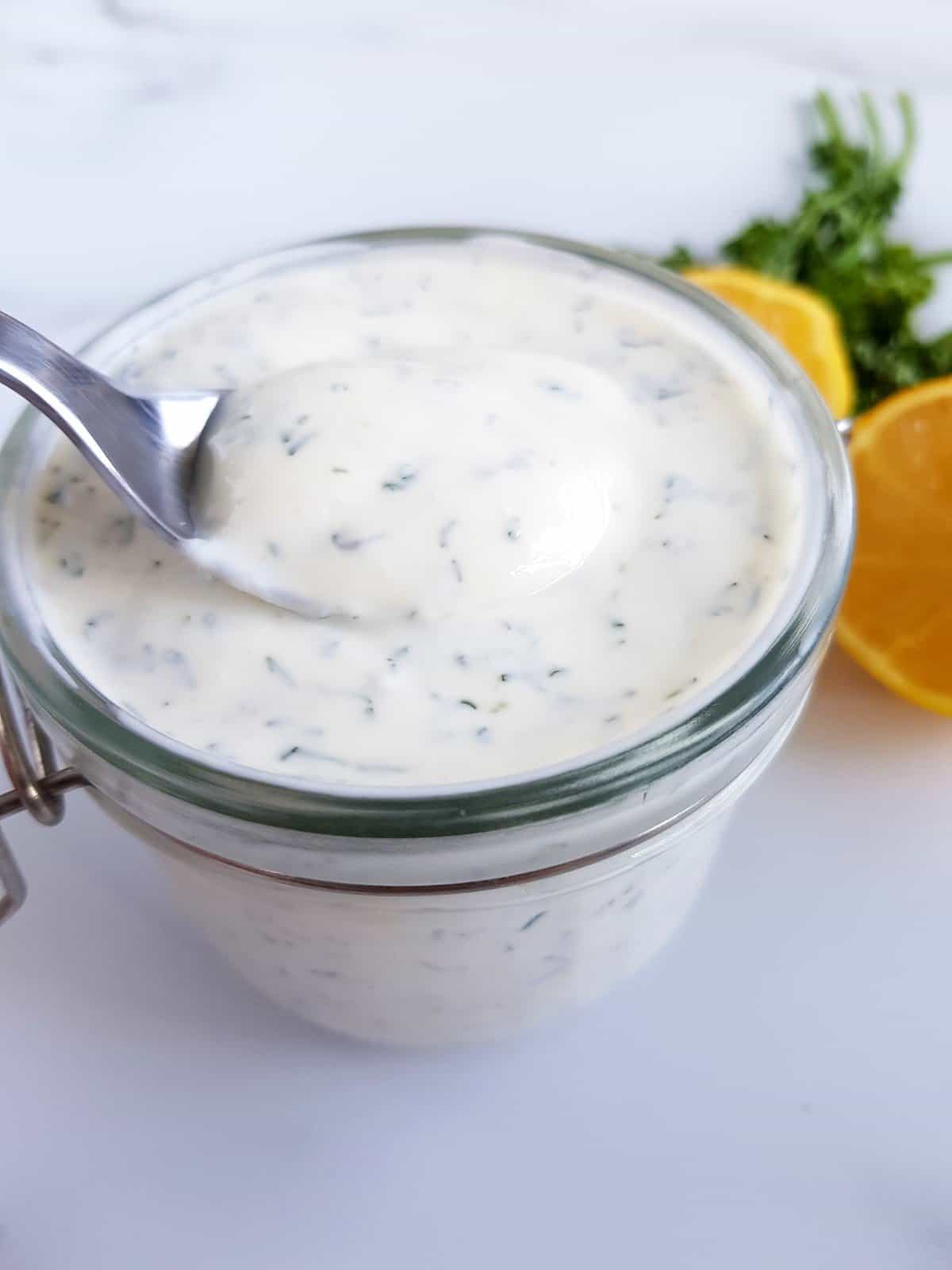 Homemade ranch in a jar with lemon and parsley on the side, a spoon being used to serve it.