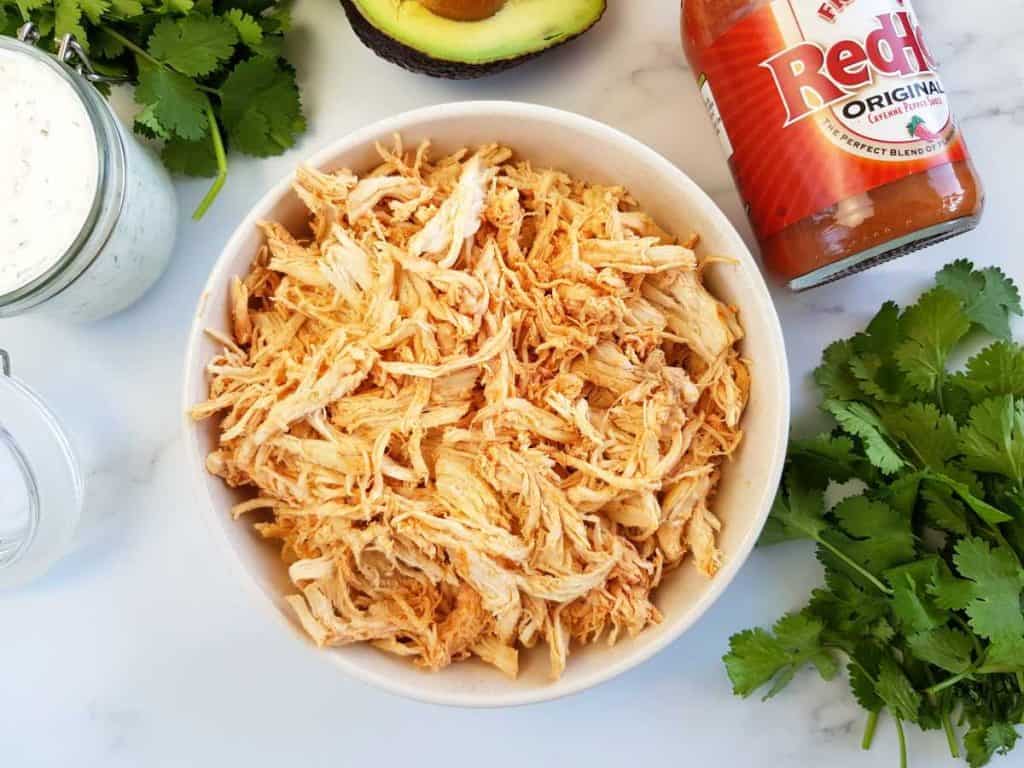 Shredded buffalo chicken in a bowl with buffalo sauce, ranch, avocado and cilantro on the side.