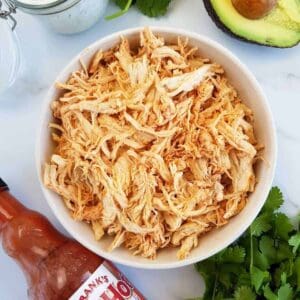 Shredded buffalo chicken in a bowl with buffalo sauce, ranch, avocado and cilantro on the side.