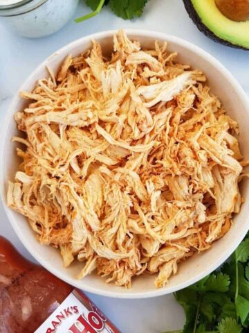 Shredded buffalo chicken in a bowl with buffalo sauce, ranch, avocado and cilantro on the side.