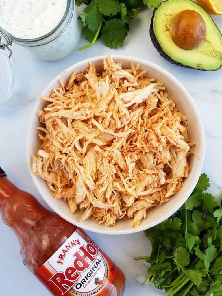 Shredded buffalo chicken in a bowl with buffalo sauce, ranch, avocado and cilantro on the side.