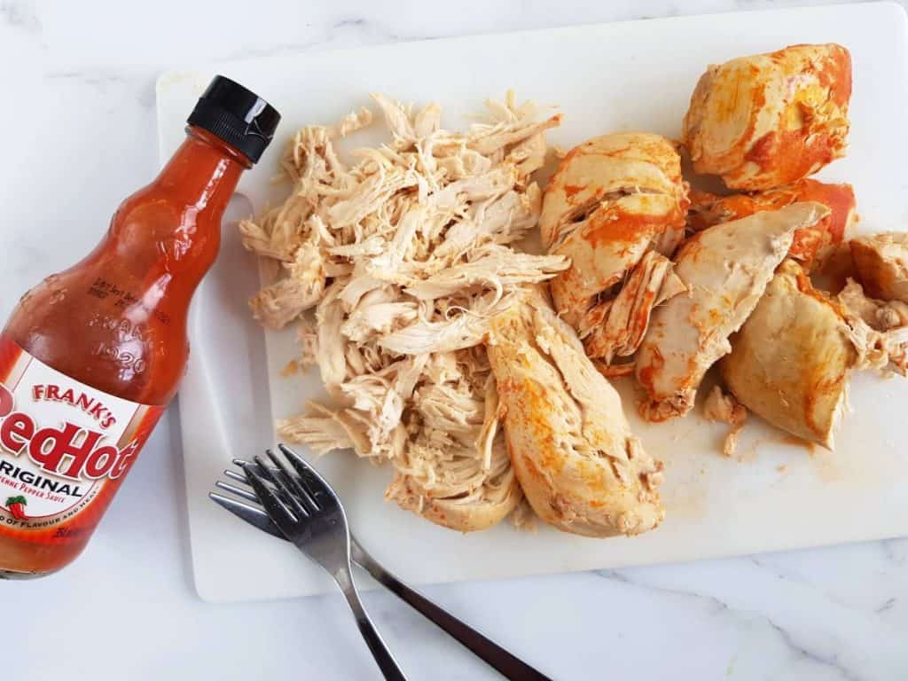 Buffalo chicken on a chopping board with two forks and buffalo sauce on the side.