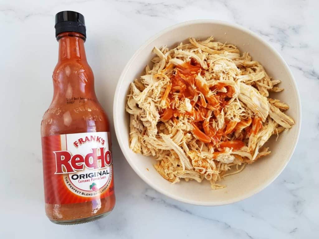 Shredded buffalo chicken in a bowl with buffalo sauce on the side.