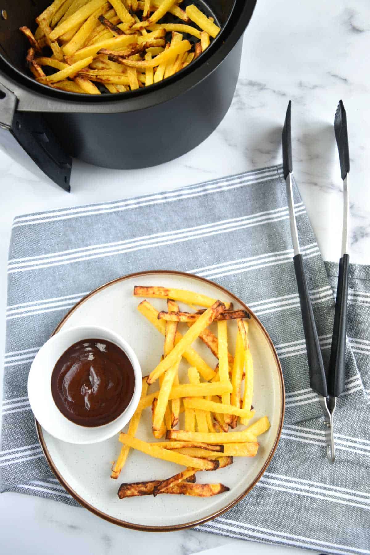 Rutabaga fries on a plate with BBQ sauce, with tongs and an air fryer next to it.