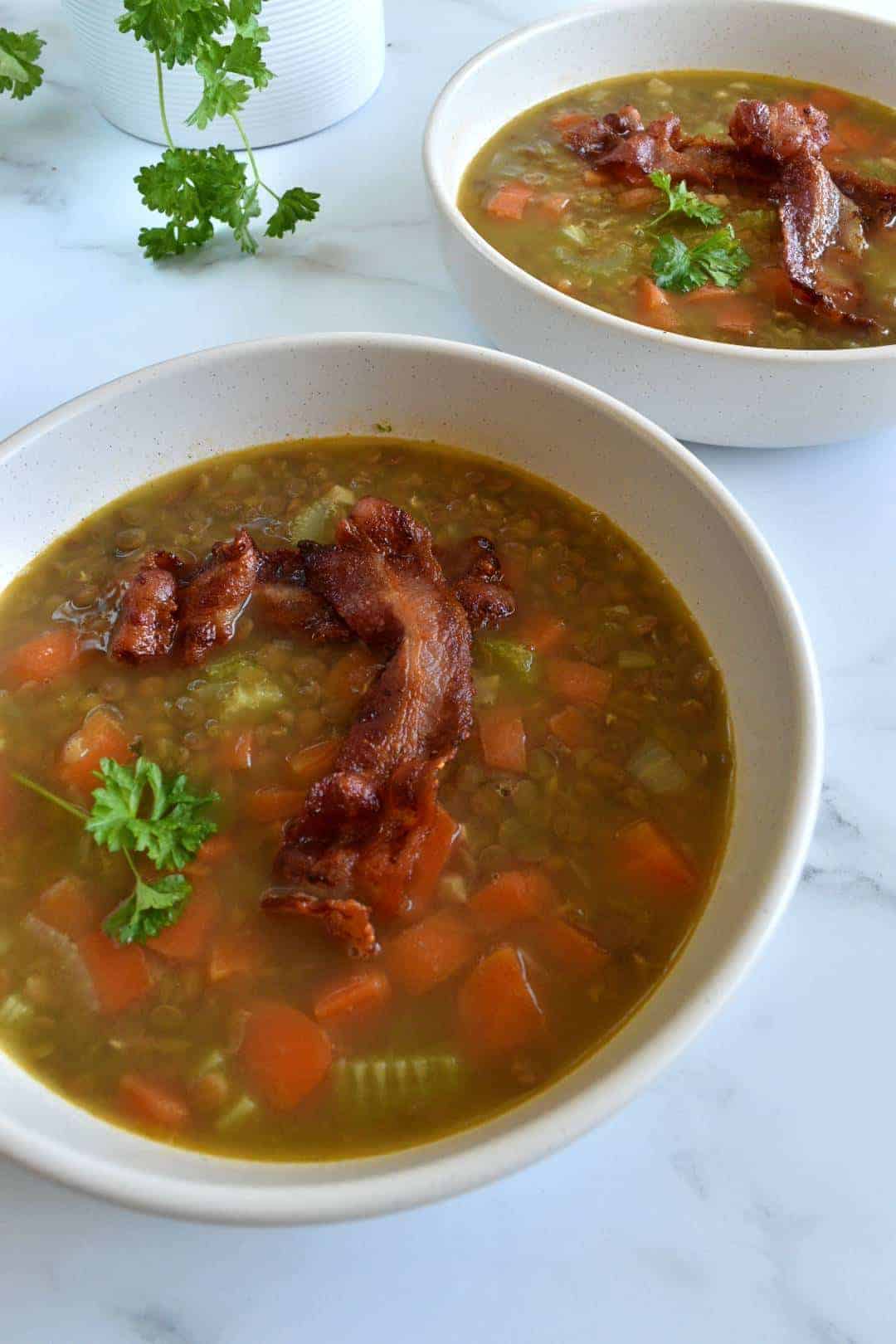 Bacon and lentil soup in white bowls on a marble table, with fresh herbs in the background.