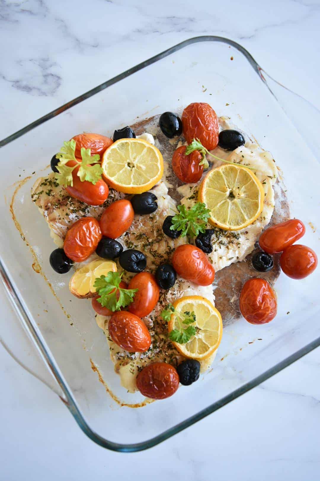 Tomatoes, olives and cod bake with fresh herbs and lemon sliced on top.