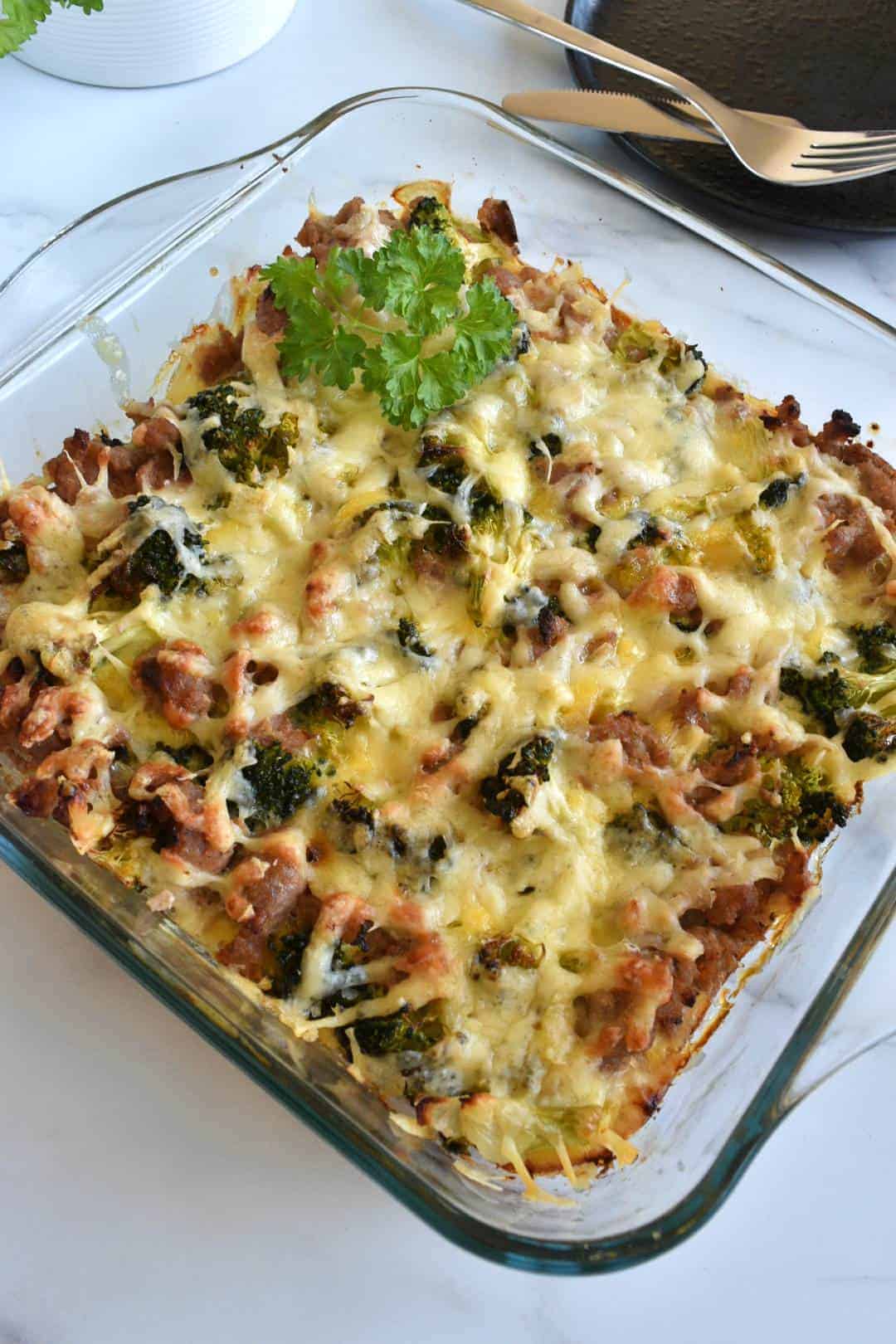 Broccoli and sausage casserole in a dish with a plate and cutlery in the background.