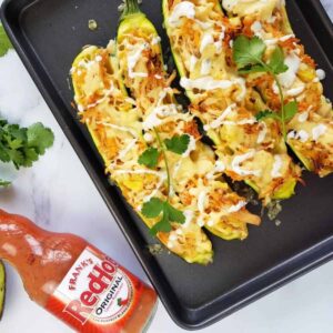 Buffalo chicken stuffed zucchini on a baking tray with a bottle of hot sauce on the side.