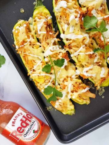 Buffalo chicken stuffed zucchini on a baking tray with a bottle of hot sauce on the side.