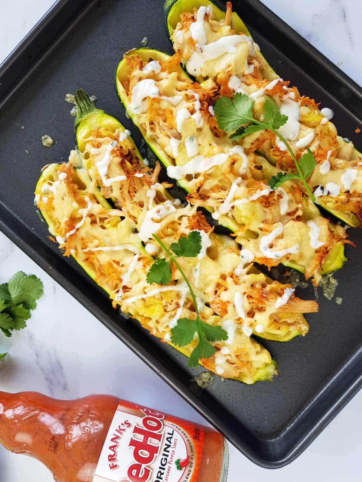 Buffalo chicken stuffed zucchini boats on a baking tray with buffalo sauce on the side.