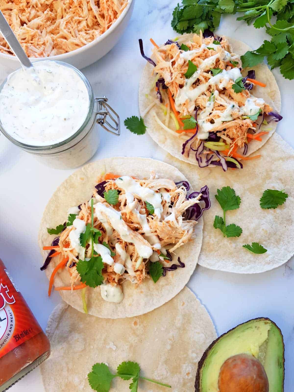 Two buffalo chicken tacos on a marble table with cilantro, ranch, avocado and buffalo chicken on the side.