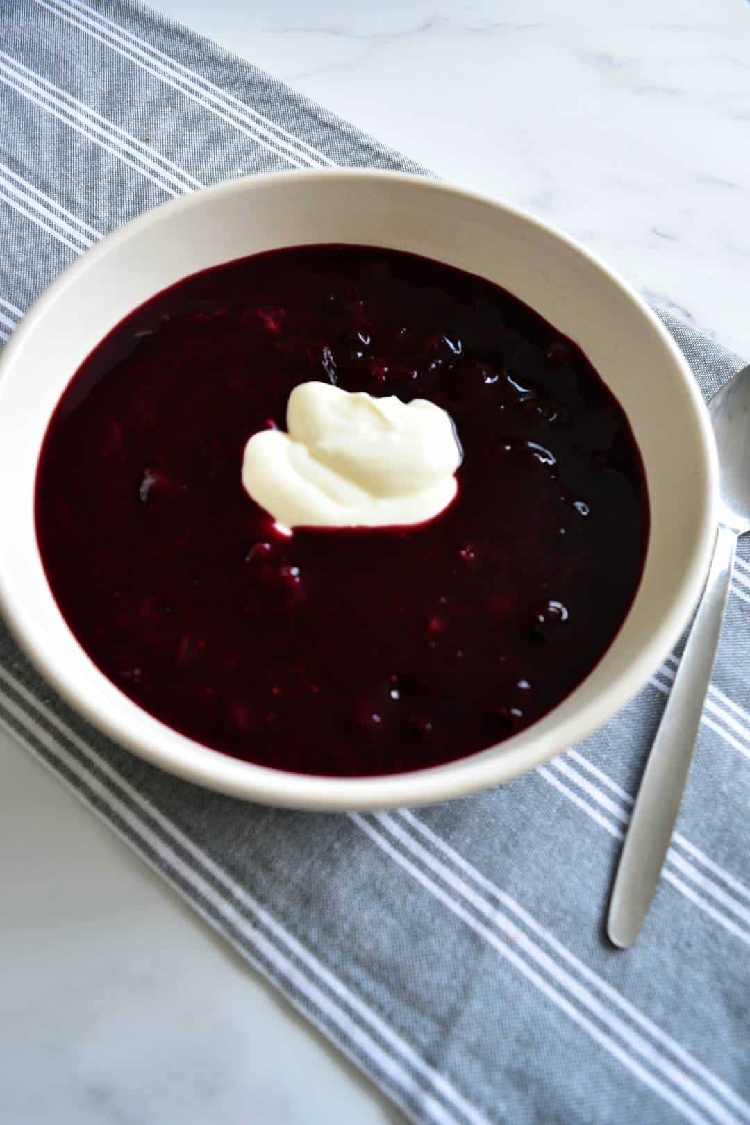 Blueberry soup topped with yogurt in a white bowl with a spoon on the side.