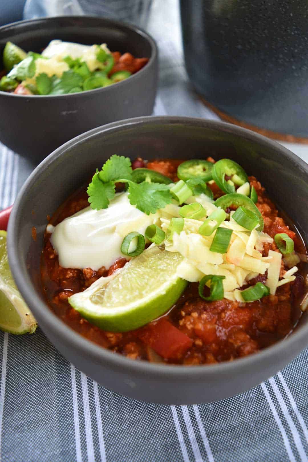 Two bowls of turkey chili topped with Greek yogurt, cheese, spring onions, limes and jalapenos.