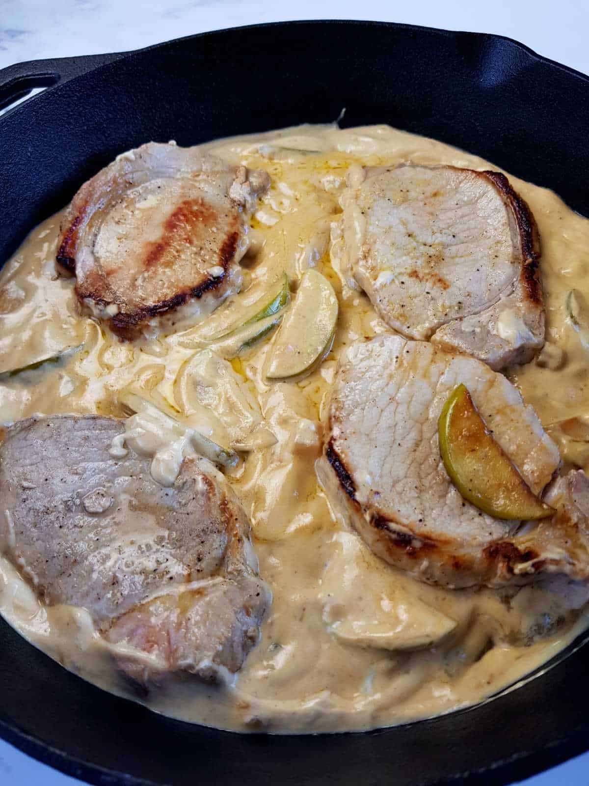 Pork chops in apple cream sauce with apple slices in a cast iron pan.