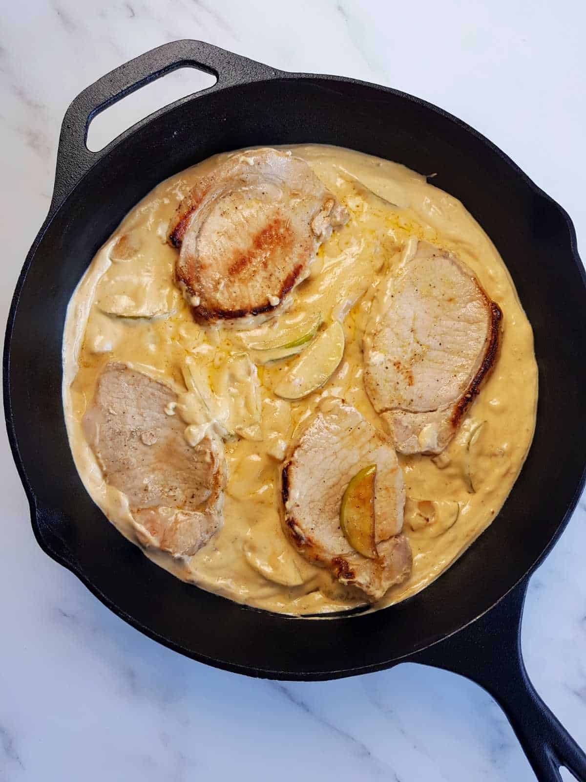 Pork chops in apple cream sauce in a cast iron skillet.