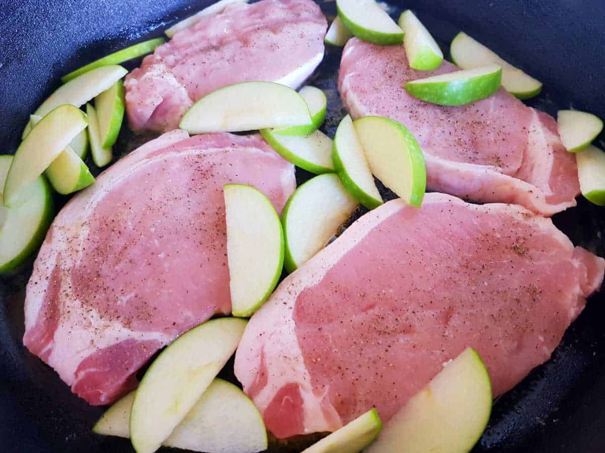 Pork chops with fresh apple slices cooking in a cast iron skillet.