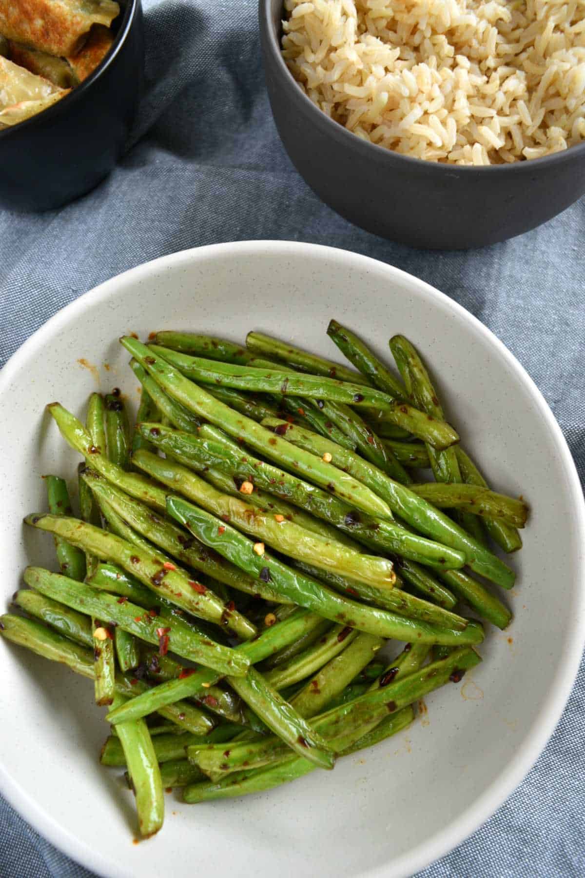 Chinese garlic green beans in a white bowl with rice and potstickers in the background.