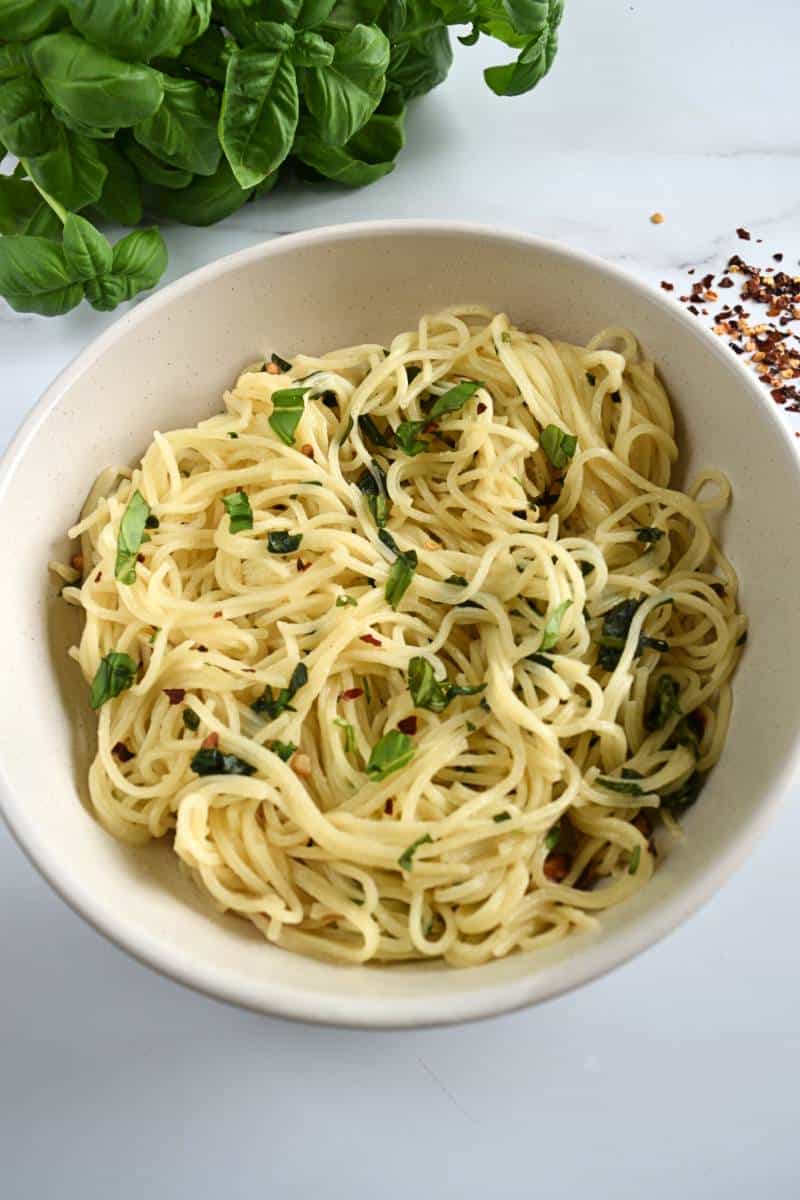 Pasta al ajo y albahaca con copos de chile en un bol blanco, con hierbas frescas al fondo.