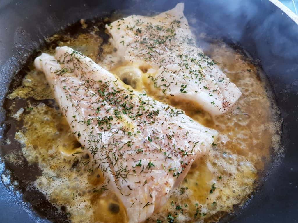 Cod cooking in a pan with butter and herbs.