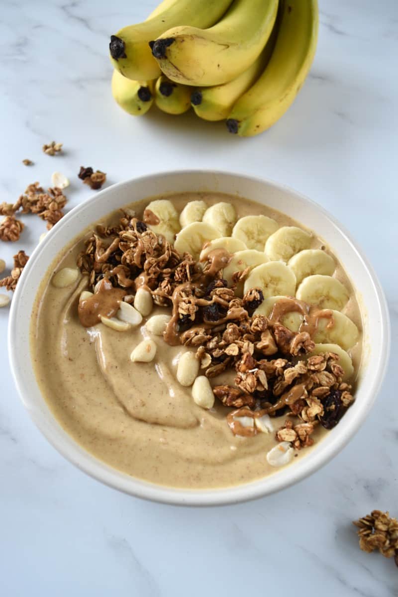 Smoothie bowl with peanut butter and raisin granola with bananas in the background.