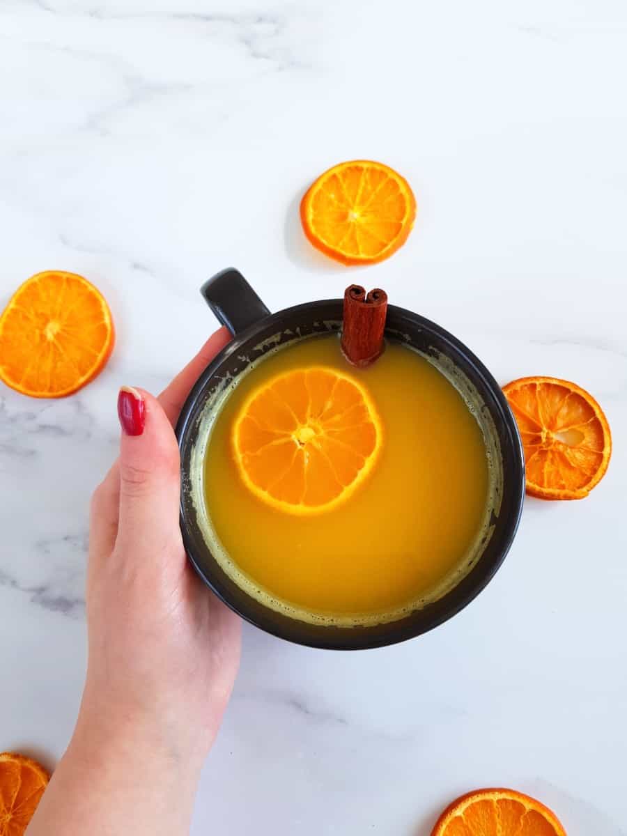 Orange juice with a cinnamon stick and orange slices in a cup, surrounded by orange slices on a marble table.