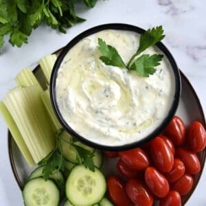 Tzatziki sauce on a plate with fresh parsley, cucumber, celery sticks and plum tomatoes.