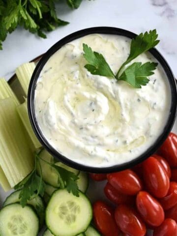 Tzatziki sauce on a plate with fresh parsley, cucumber, celery sticks and plum tomatoes.